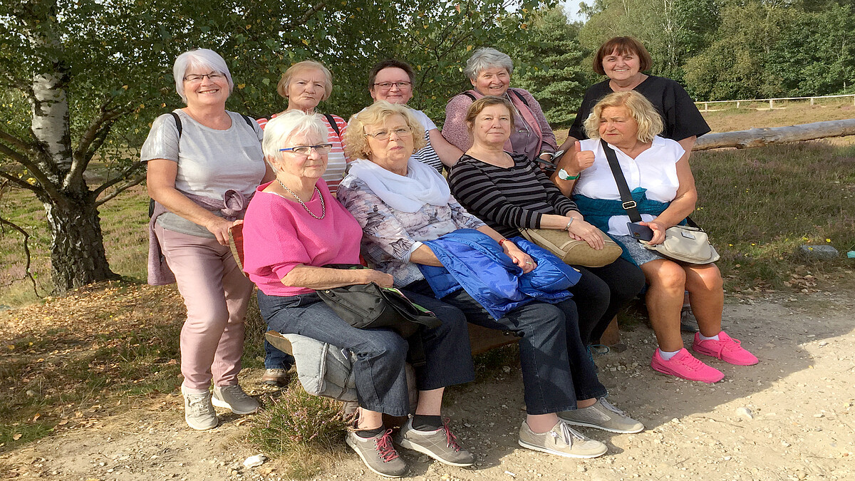 Acht ältere Frauen stehen und sitzen rund um eine Bank in einer Heidelandschaft.