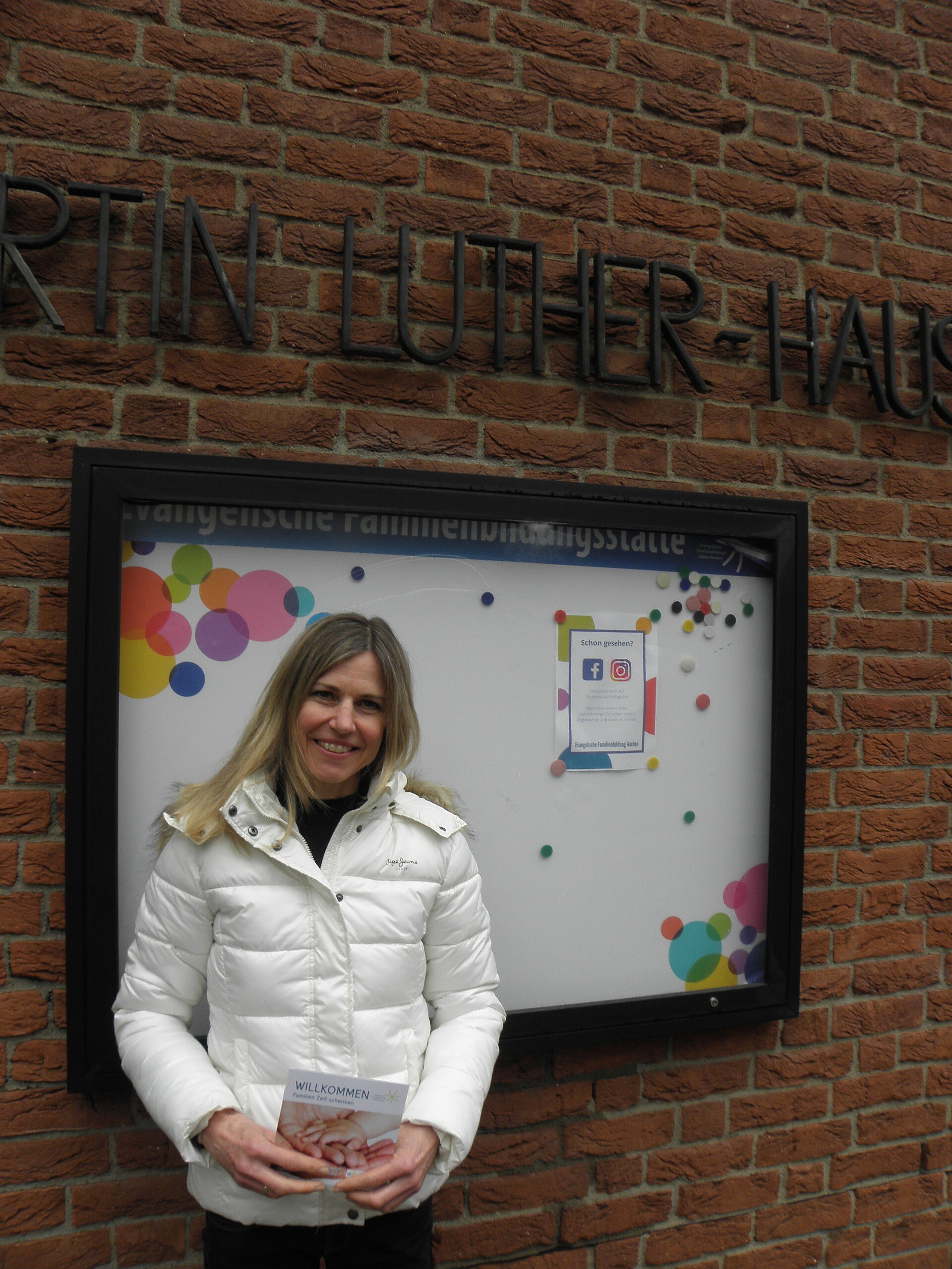 Junge Frau in weißer Jacke steht vor einer Infotafel der Evangelischen Familienbildungsstätte Aachen.
